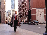 Video Clip - Man with a brown trench coat and a brief case walks down a street and waits at a street light